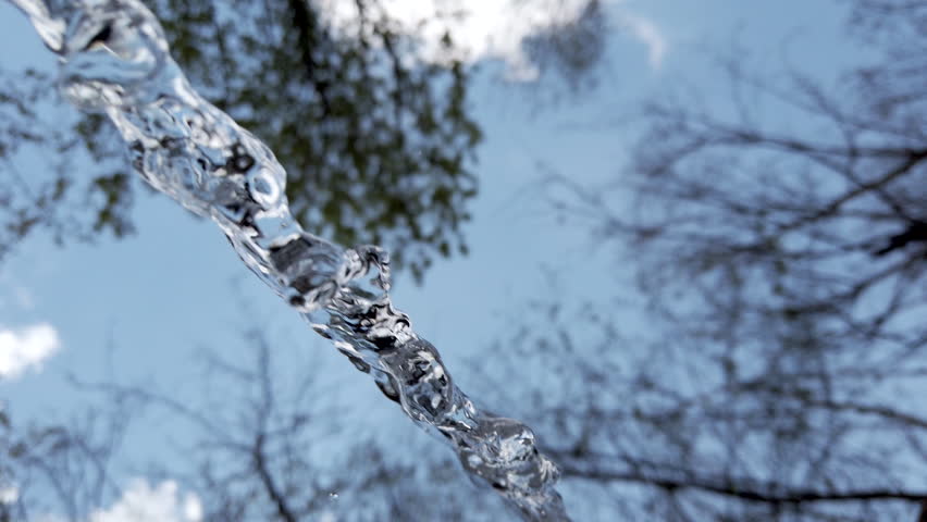 Water streams out from a hose, creating clear shapes as it flows. Above, branches stretch across the bright blue sky. This takes place in a green park on a warm afternoon.