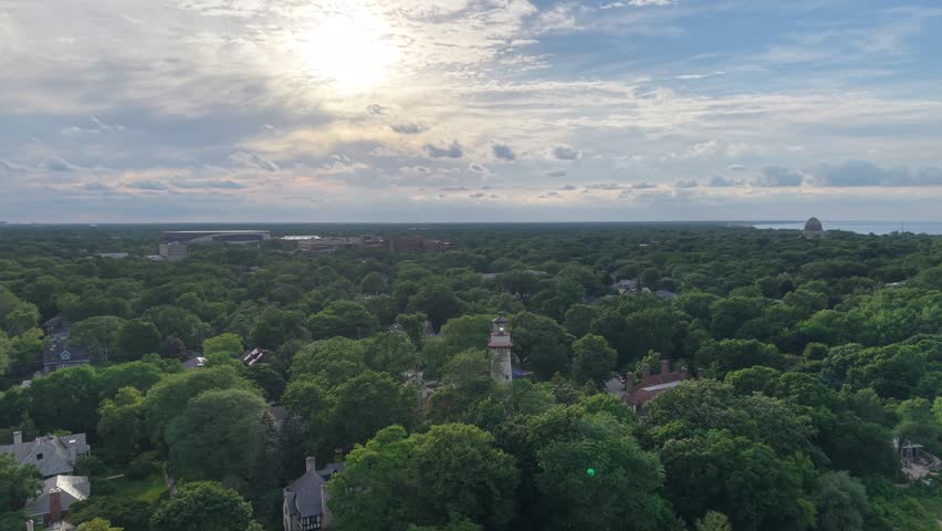 Golden-hour aerial of the Grosse Point Lighthouse in Evanston, Illinois, set among lush summer treetops along the Lake Michigan shoreline, with the Bahá