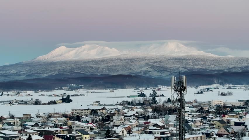 This winter landscape photograph captures a small snow-covered Higashikawa town nestled in a broad valley beneath a majestic mountain range 大雪山系 Daisetsuzan Volcanic Group. The mountains in the background are blanketed in fresh snow, their peaks glowing softly under diffused daylight and a pastel-colored sky. In the foreground, houses, roads, and open fields are covered with a thick layer of snow, creating a peaceful rural atmosphere.