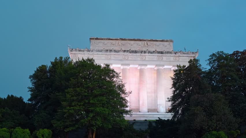 Lincoln Memorial illuminated at twilight, Washington, D.C. Warm pink light on marble columns framed by dark silhouetted trees against deep blue sky.