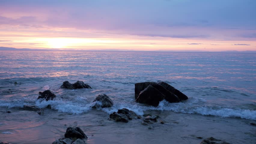 Static handheld shot of dark rocks on the shore of Lake Baikal during sunset. Gentle waves wash over the stones under a beautiful pink and purple evening sky.