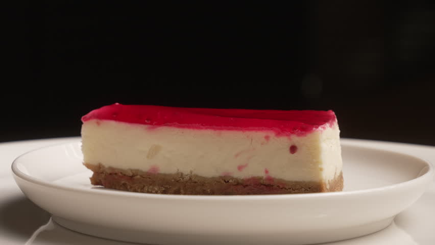 Slice of sweet strawberry cheesecake with red jelly topping rotating on a white plate, fa01