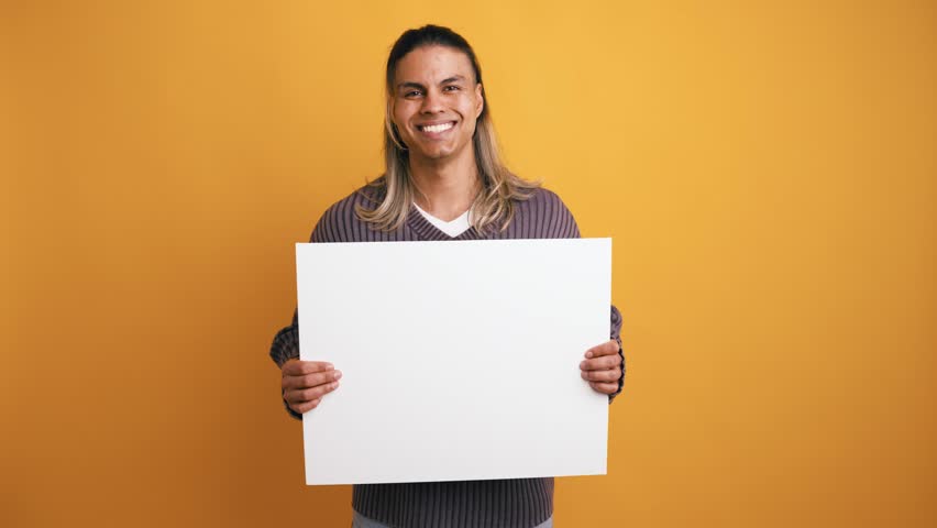 Handsome young man with long hair holding a blank white poster. Casual person standing over a yellow background with a mockup sign
