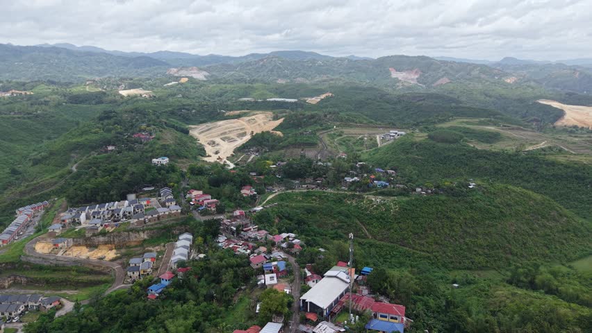 Aerial drone view of a mountainous landscape featuring residential communities, rolling green hills, and ongoing land development activities in the Philippines. The footage showcases a mix of rural settlements, infrastructure expansion, and natural scenery under a cloudy sky.