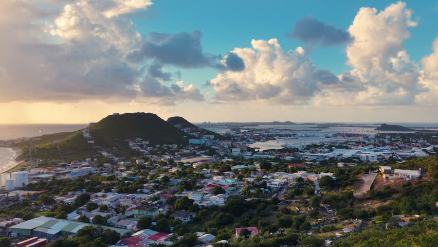 Aerial view of Cole Bay town and hills on the Dutch side of Sint Maarten island. Panoramic coastal landscape of the Caribbean destination under a cloudy sky. Buildings and residential houses spread across the valley near the waterfront. Simpson Bay Lagoon with boats visible in the background under daylight.