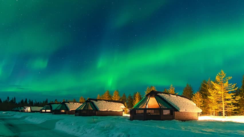 Detailed northern lights shimmering over winter cabins in arctic forest landscape. Vibrant green aurora borealis motion timelapse with starry night sky and snowy ground scenery.