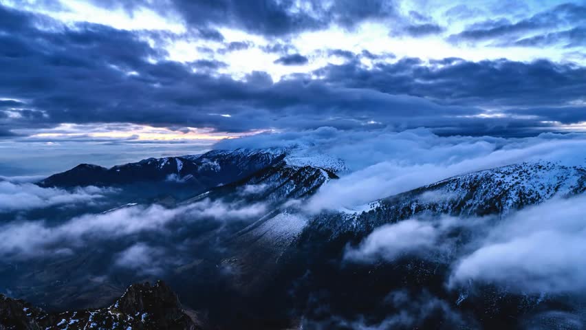 Aerial view of majestic snow covered mountain peaks shrouded in mist and fog. Dramatic clouds drift across the sky at sunrise.