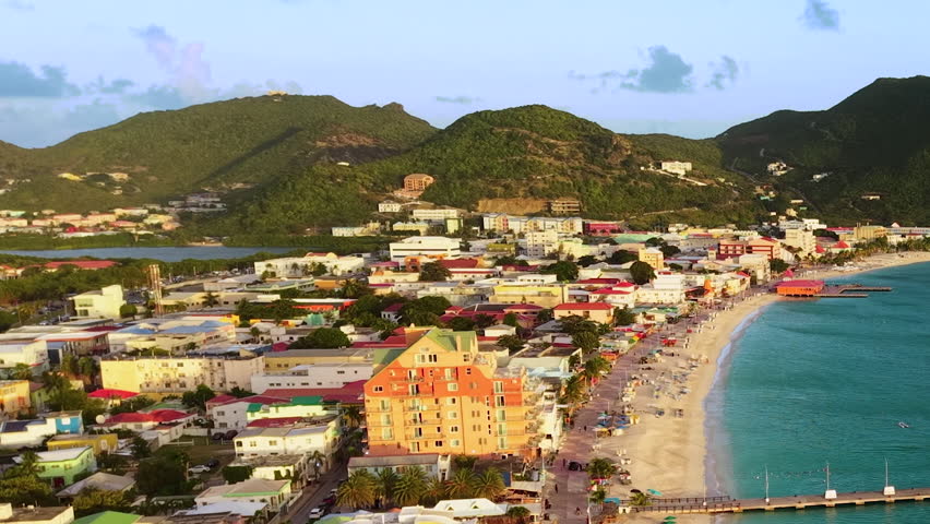 Aerial view of Great Bay beach and Philipsburg harbor on the Dutch side of Saint Martin island. Blue Caribbean sea water features scattered sailboats and a large cruise ship docked in the distance. Green lush hills border the coastal town with its sandy shoreline and waterfront buildings. Drone footage captures the tropical paradise landscape under a partly cloudy sky.