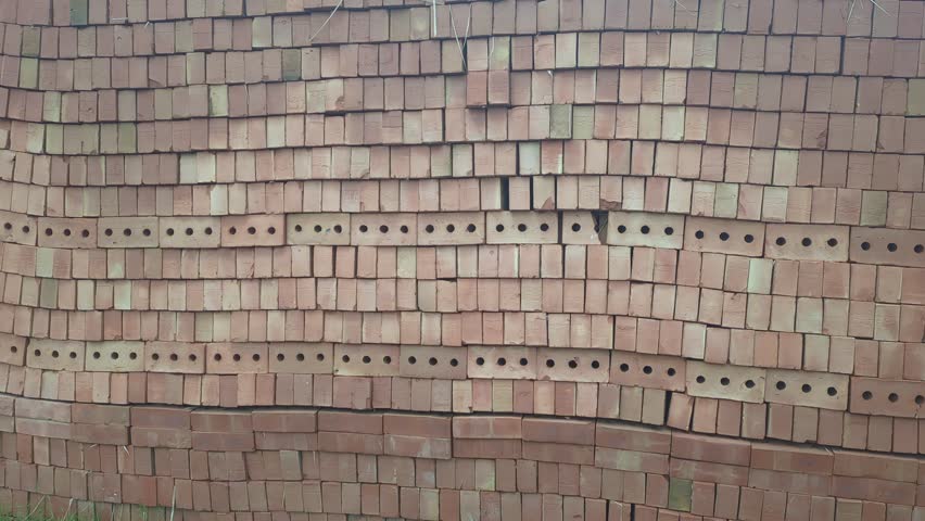 A massive stack of red bricks, meticulously arranged in a neat wall. Some of the rows feature hollow bricks with uniform round holes, adding texture to the surface. The camera remains mostly static, highlighting the clean, grid-like pattern and the subtle variations in brick tones.