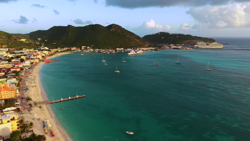 Aerial view of Great Bay beach and Philipsburg harbor on the Dutch side of Saint Martin island. Blue Caribbean sea water features scattered sailboats and a large cruise ship docked in the distance. Green lush hills border the coastal town with its sandy shoreline and waterfront buildings. Drone footage captures the tropical paradise landscape under a partly cloudy sky.
