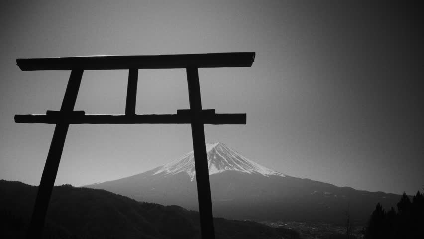 Black and white view of a traditional Japanese torii gate framing snow-capped Mount Fuji above forested hills in the Japanese countryside
