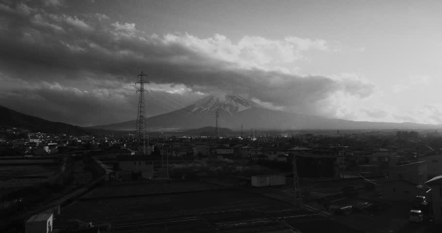 Black and white aerial drone shot rising above residential neighborhoods of Fujiyoshida with Mount Fuji partially shrouded by dramatic clouds and evening sunlight breaking through the sky in Yamanashi Japan