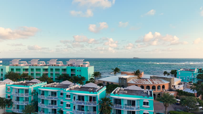 Aerial view of a coastal tropical resort on the eastern coast of Saint Martin island in the Caribbean Sea. Modern hotel buildings feature distinct white and blue architectural designs near the water. This luxury vacation destination is located in the Dutch side of the island. Turquoise ocean waves splash against the shoreline under a partly cloudy sky.