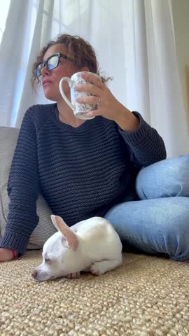 Woman in casual clothes sitting on a comfortable sofa, drinking from a mug and looking thoughtfully out the window while her little white chihuahua dog sleeps peacefully beside her