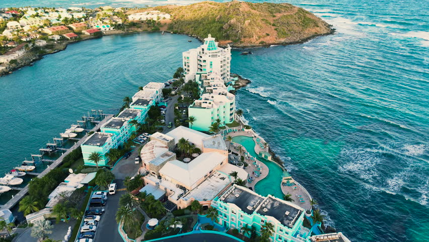 Drone view of the luxury beach resort situated on a peninsula. The modern hotel buildings stand on the coastline facing the Caribbean Sea. This location featuring a large curved swimming pool is situated on the Dutch side of Saint Martin island. Golden hour sunlight illuminates the resort architecture, sandy shoreline and turquoise ocean water.