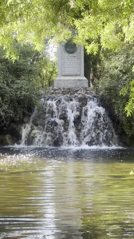 Madrid, Spain - June 02, 2026: Waterfall and pond inside a public park surrounded by trees, with a stone monument in the background.