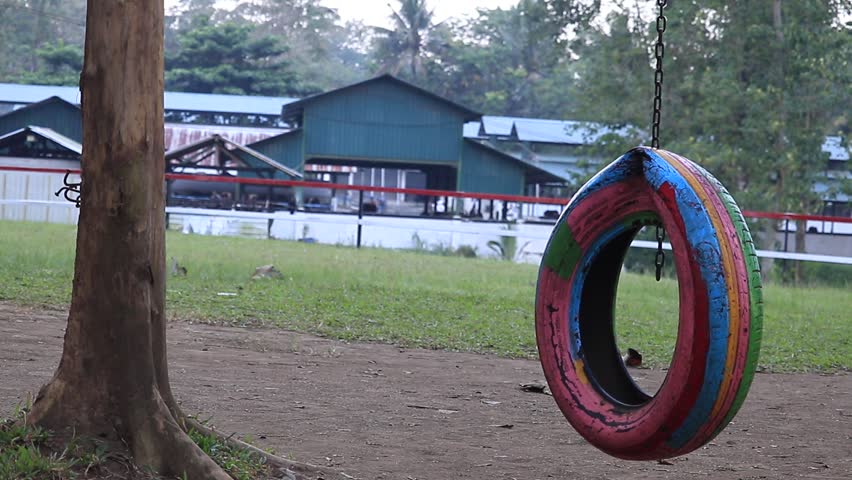 A vibrant tire swing suspended from a tree in a park with buildings and greenery in the background on a cloudy day