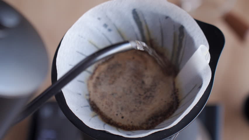 Hot water pours over ground coffee in a paper filter, creating coffee bloom in a pour over dripper. Process of person preparing drip coffee at home