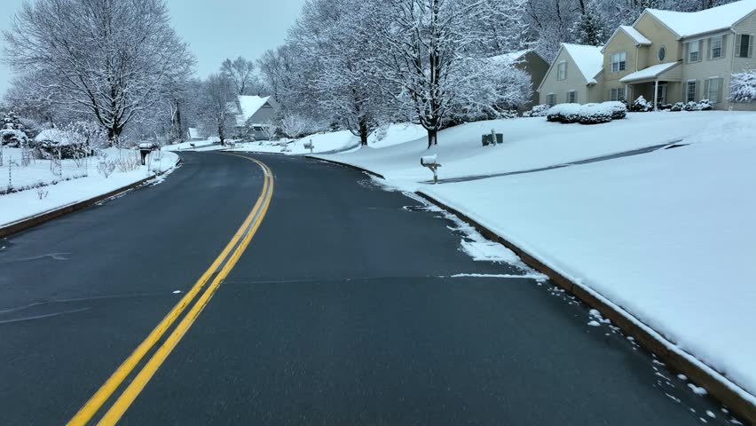 Slow camera movement traveling along paved road through suburban neighborhood during winter season featuring snow covered trees houses and residential lawns under overcast sky scenery.