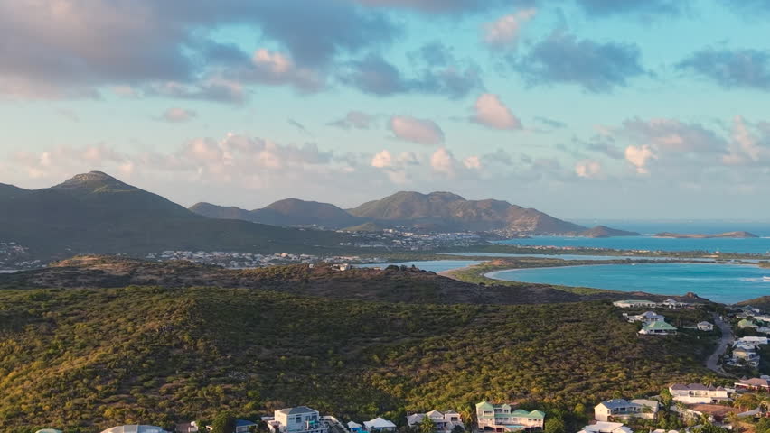 Drone aerial view of Oyster Pond bay on the border of Sint Maarten and Saint Martin. Tropical island landscape features luxury coastal houses and boats anchored in the calm harbor. Green hills and blue ocean waters stretch toward the horizon under a cloudy sky. Scenic destination on the Caribbean coast perfect for travel, real estate, and vacation themes.