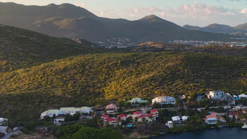 Drone aerial view of Oyster Pond bay on the border of Sint Maarten and Saint Martin. Tropical island landscape features luxury coastal houses and boats anchored in the calm harbor. Green hills and blue ocean waters stretch toward the horizon under a cloudy sky. Scenic destination on the Caribbean coast perfect for travel, real estate, and vacation themes.