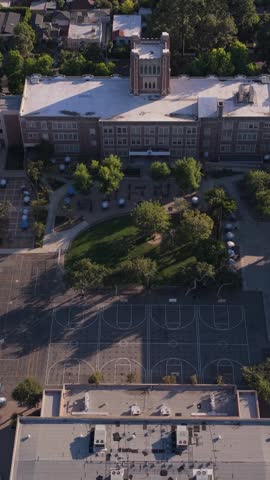 Top down orbit shot showcasing historic brick school building with clock tower, playground, basketball court in residential neighborhood of Los Angeles, California