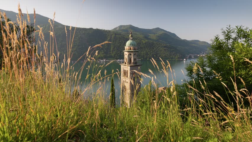Ancient church bell tower in Morcote village overlooking the calm lake water. Stabilizer shot of historical stone architecture in Ticino canton of Switzerland. Morcote landmark in Switzerland