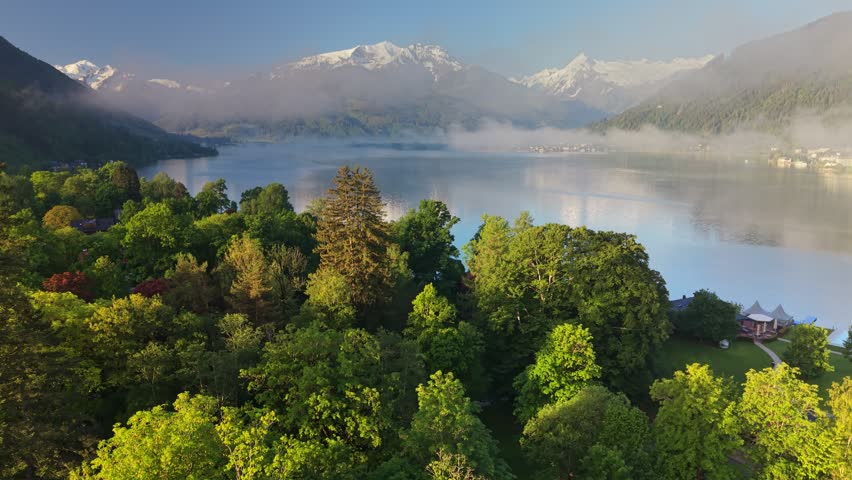 Sunrise over Zell am See mountain lake with green trees, misty water and snowy alpine peaks in Austria. Scenic aerial view of Zell am See region at sunrise in spring. Snowy Austrian Alps