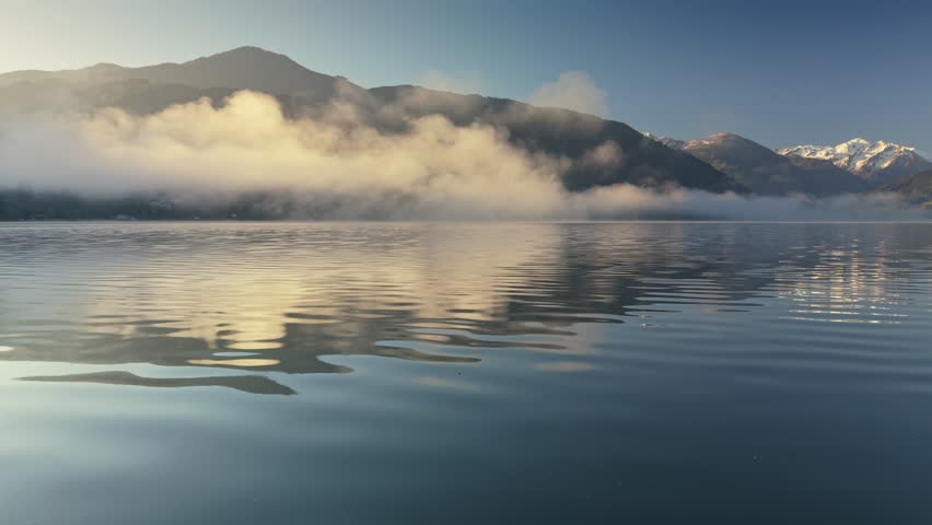 Tranquil lake morning fog in Zell am See with majestic snow covered mountains in the background. Low angle drone flyover above calm water ripples in Austria. Austrian Alps reflecting in water