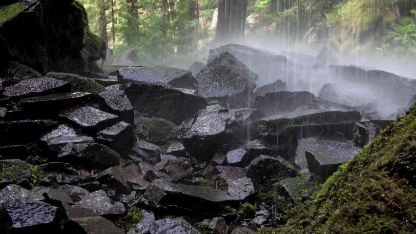 Wet dark rocks at the base of a misty waterfall in a lush green forest