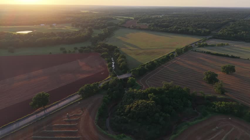 Drone orbit above harvested hay fields and a winding creek at sunset in Oklahoma featuring round bales, farmland, rural roads, and early summer agricultural scenery.