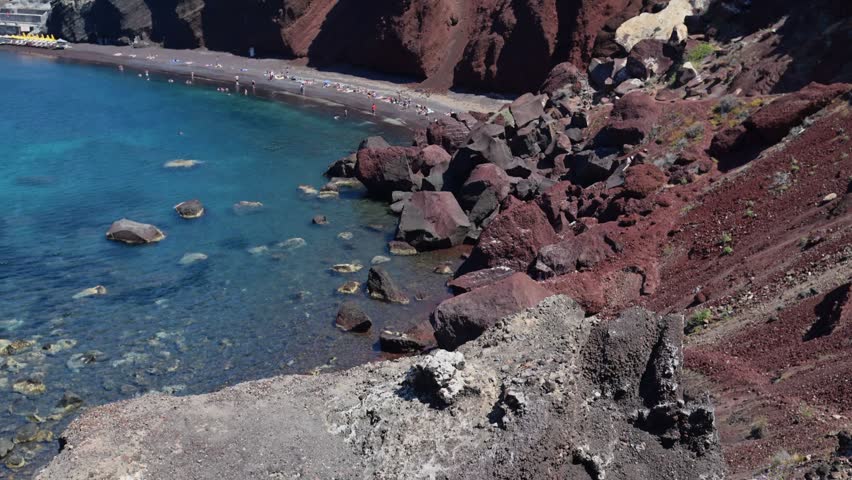 Aegean cove with clear turquoise water, dark shoreline, and dramatic red lava cliffs on Santorini, Greece.