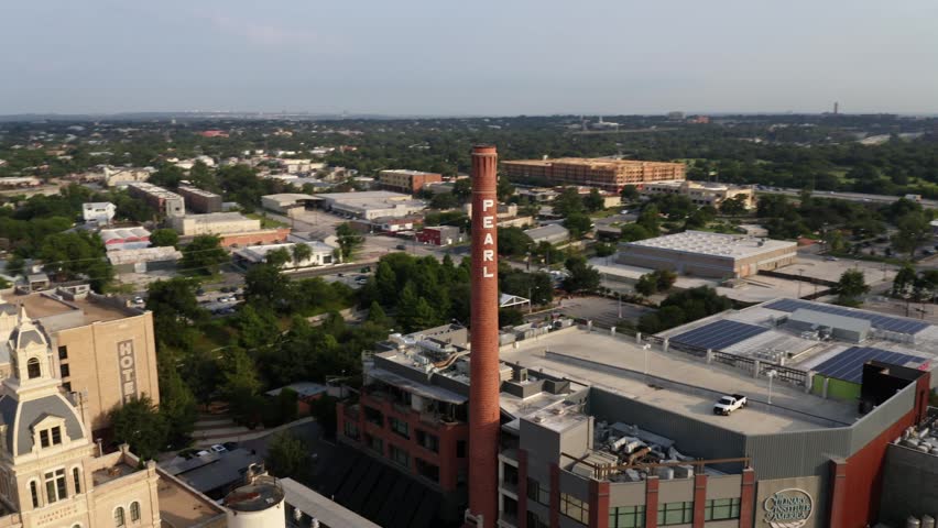 Aerial drone footage capturing iconic pearl brewery landmark with tall brick smokestack architecture and surrounding city skyline scenery during golden hour light in texas urban environment.