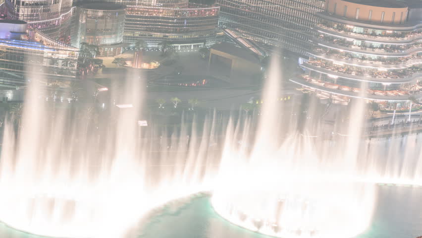 Dubai Fountain night timelapse in Downtown Dubai. Aerial view showing circular fountain with illuminated pathways and dark water. Water show captured from a high angle rooftop perspective.