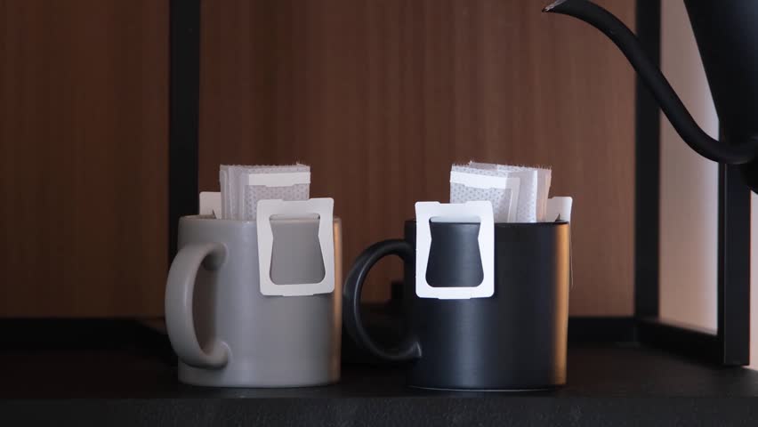 tea and coffee set for self-service on wood shelf in luxury hotel room. Coffee drip bags on cups in hotel room and kettle for alternative coffee brewing. Female hand pouring hot water into cups