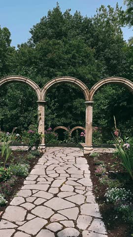 A stone garden path leads beneath tall arched columns surrounded by green trees, leafy plants, and small colorful flowers. The setting includes textured stone, classic garden architecture, bright sky, and a peaceful walking route through a landscaped outdoor space.
