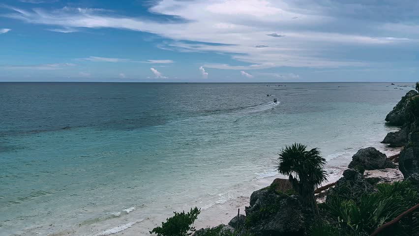 A wide Cancun Tulum Mexico ocean view with blue water stretching toward the horizon beneath a bright sky with soft clouds. A small motorboat moves across the sea while a sailboat appears in the distance, creating a calm coastal travel setting.
