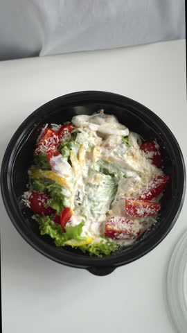 Person mixing fresh Caesar salad with chicken and parmesan cheese in a takeaway bowl with a fork on a white table