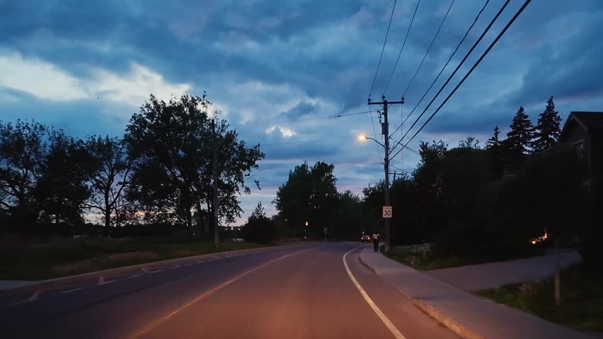A scenic riverside roadway curves through Montreal, Quebec, Canada during sunset. Colorful evening skies, dramatic clouds, and distant residential buildings create a calm and atmospheric urban driving scene.