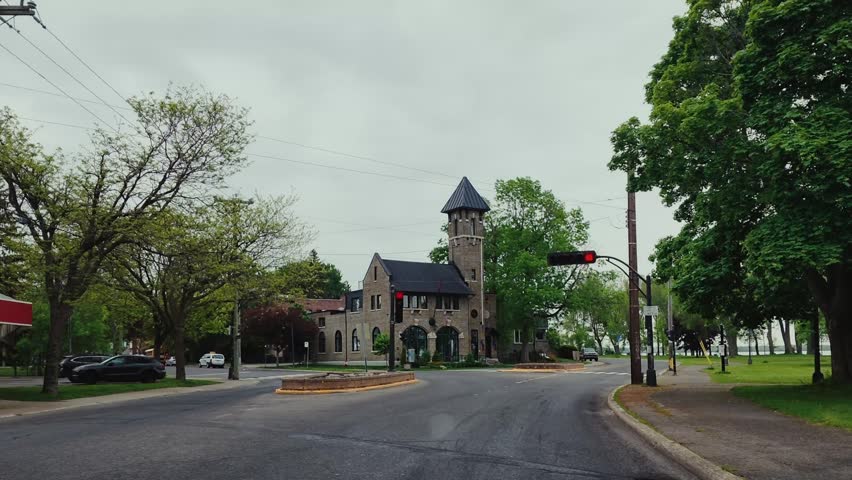 POV driving footage approaching a flashing red traffic light near a waterfront road in Montreal, Quebec, Canada. A unique riverside building, open parkland, and quiet streets create an atmospheric city landscape.