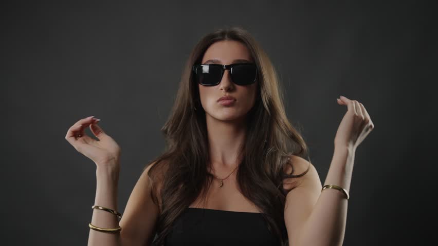 Female model posing in sunglasses, embodying beauty and fashion concepts in a studio setting. The woman with long brunette hair and bracelets presents a confident and elegant style on a dark backgroun