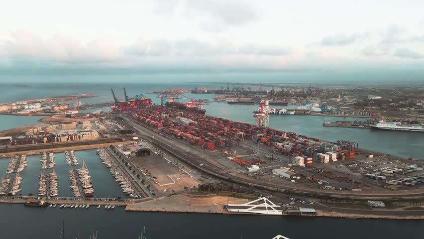 Aerial view shows a busy container port. Red shipping containers line the docks. Cranes and ships fill the harbor area. Curved waterway flows through the port