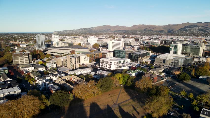 Dynamic aerial footage showcasing a sprawling christchurch city with diverse architecture, lush trees, and distant mountains under a bright, sunny sky, creating a picturesque urban landscape.
