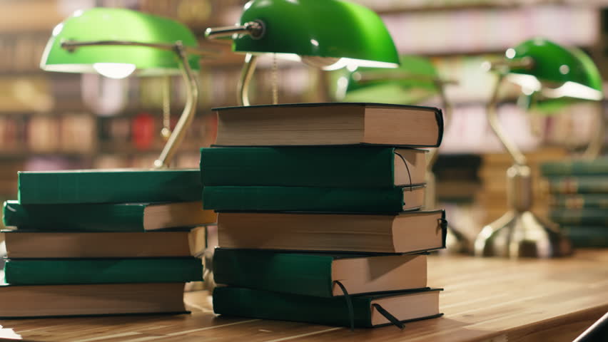 Close-up view shows stacked books on desk in quiet university library with warm lighting and green lamp, educational environment with bookshelves and study materials, empty library study space.