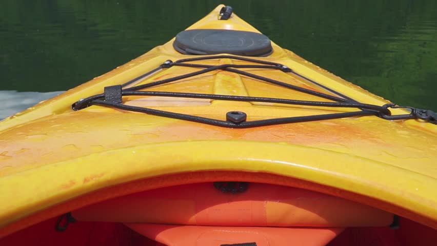 The bow of a yellow kayak slices through still, reflective lake waters, surrounded by lush green hills. Ideal for adventure content or outdoor lifestyle themes.
