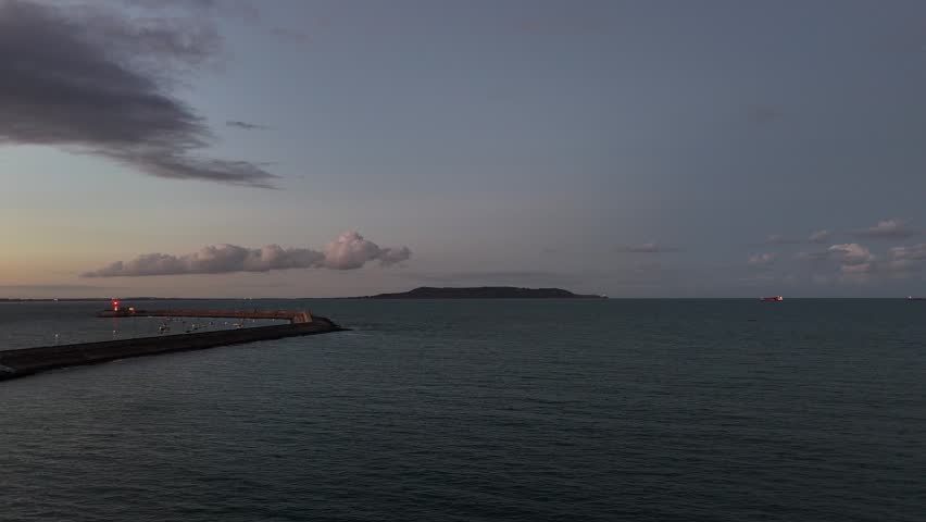 Cinematic aerial drone footage pulling back from a solitary sailing boat in Dublin Bay towards the South Dublin coastline. As the drone retreats, expansive views of Dún Laoghaire Harbour and the shoreline unfold, culminating in the distinctive silhouette of the Roger Casement statue overlooking the coast. Filmed on a calm evening with soft twilight light, capturing the maritime character and coastal beauty of Dublin
