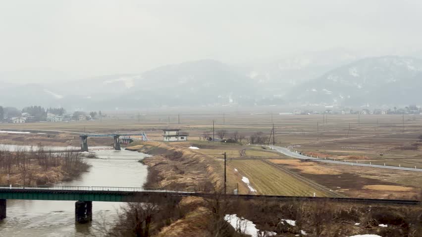 A tracking shot from a train window reveals a snowy Japanese valley under overcast skies