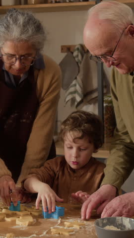 Vertical video of senior Caucasian grandparents helping child grandson cut star shapes out of cookie dough on wooden table, perfect for family lifestyle commercials and holiday baking promotions