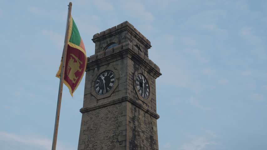 Low angle slow motion shot of the Sri Lankan flag waving beside Galle Fort Clock Tower, showing historic stone architecture and clock faces against a pale blue sky.
