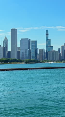 Tall buildings rise above water in a city with a clear blue sky. Boats are visible on the lake, and green spaces line the waterfront.